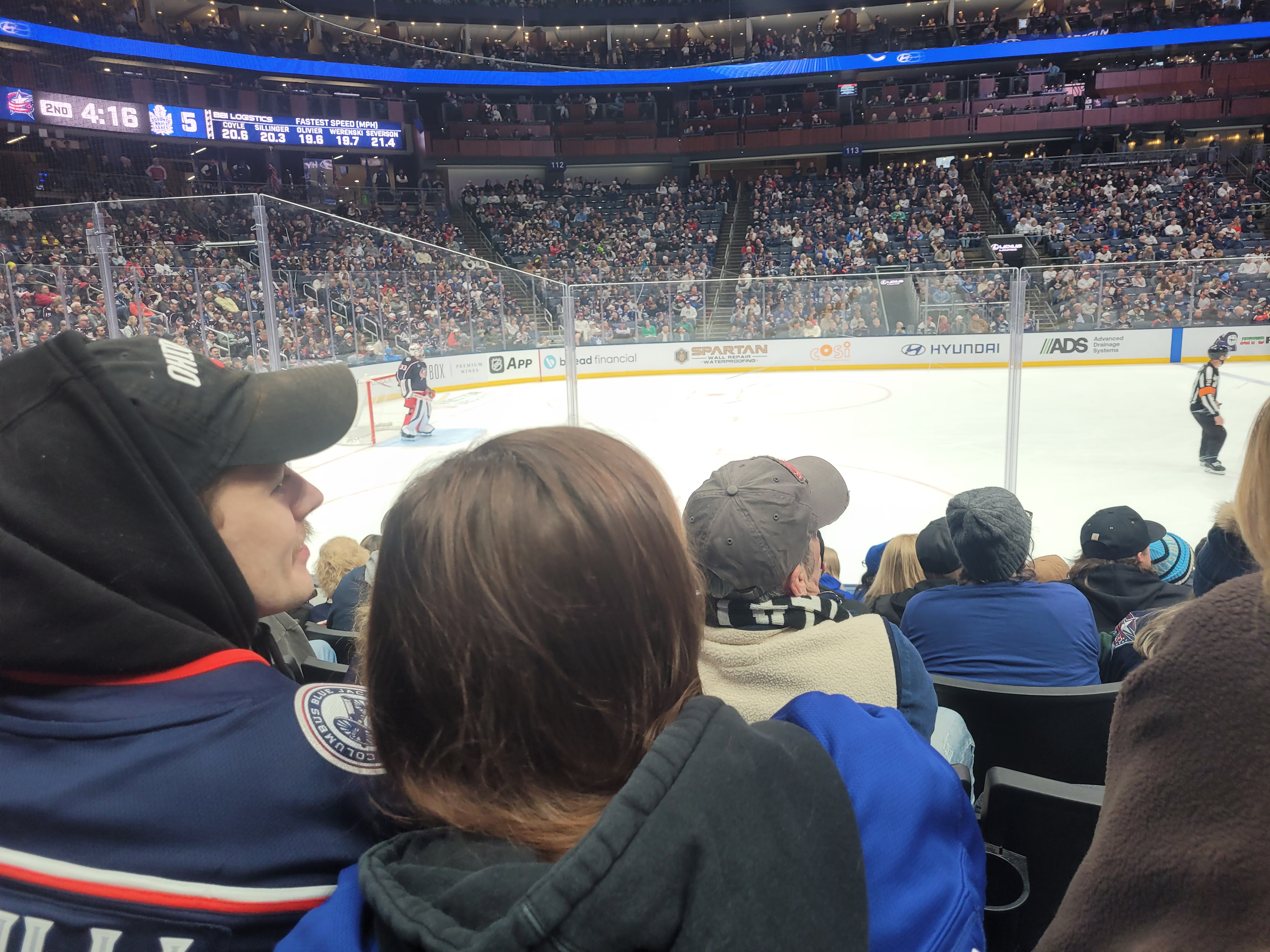 View from the lower bowl at Nationwide Arena during the second period of the Blue Jackets game against the Maple Leafs, with the Columbus goalie in net, fans in Blue Jackets sweaters in the foreground, and the scoreboard showing 4:16 left in the period
